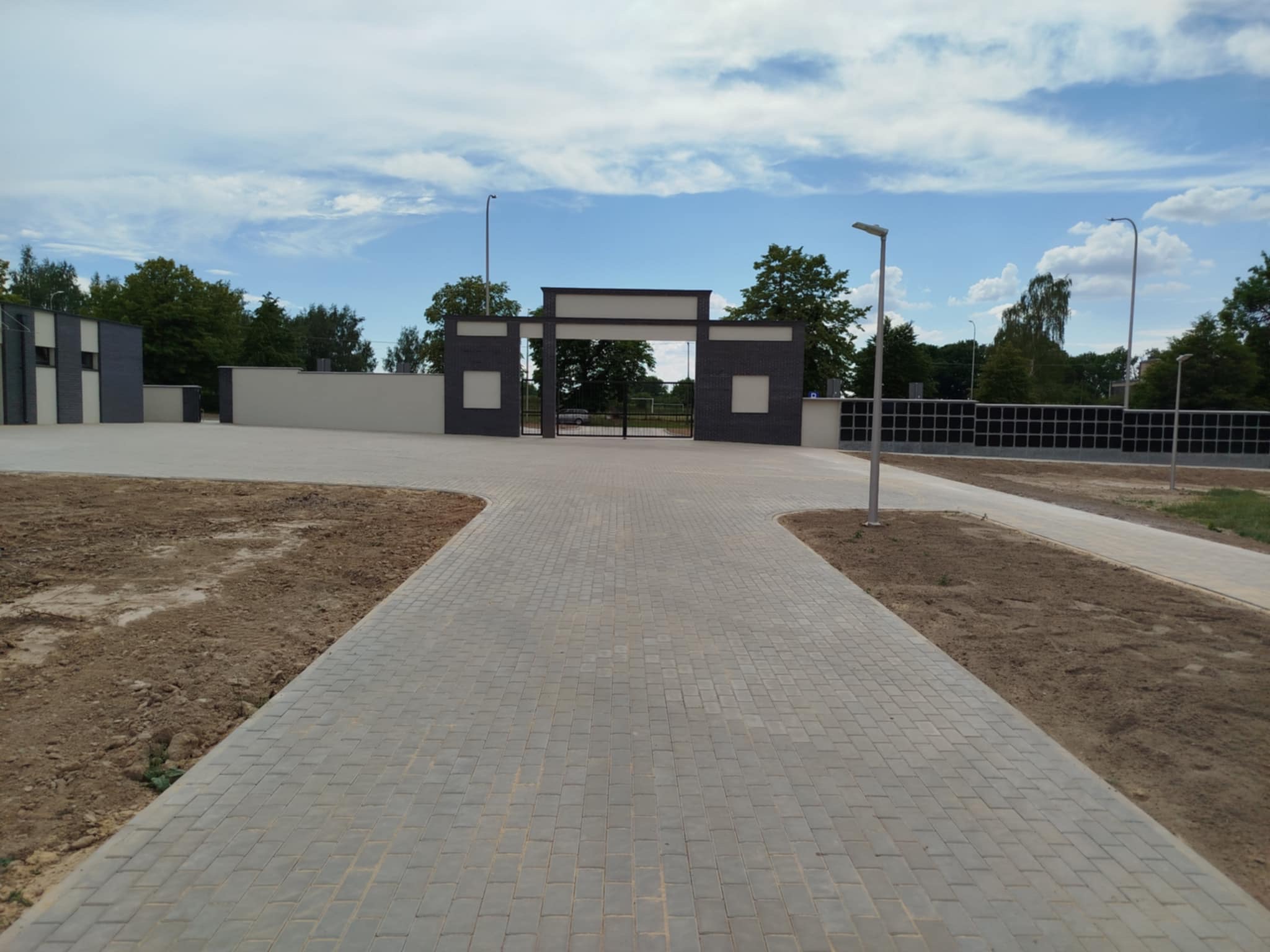Ułożona kostka brukowa tworząca ścieżkę prowadzącą do bramy z elementami murowanymi. Widoczne lampy oświetleniowe i fragmenty terenu przygotowanego pod nasadzenia.