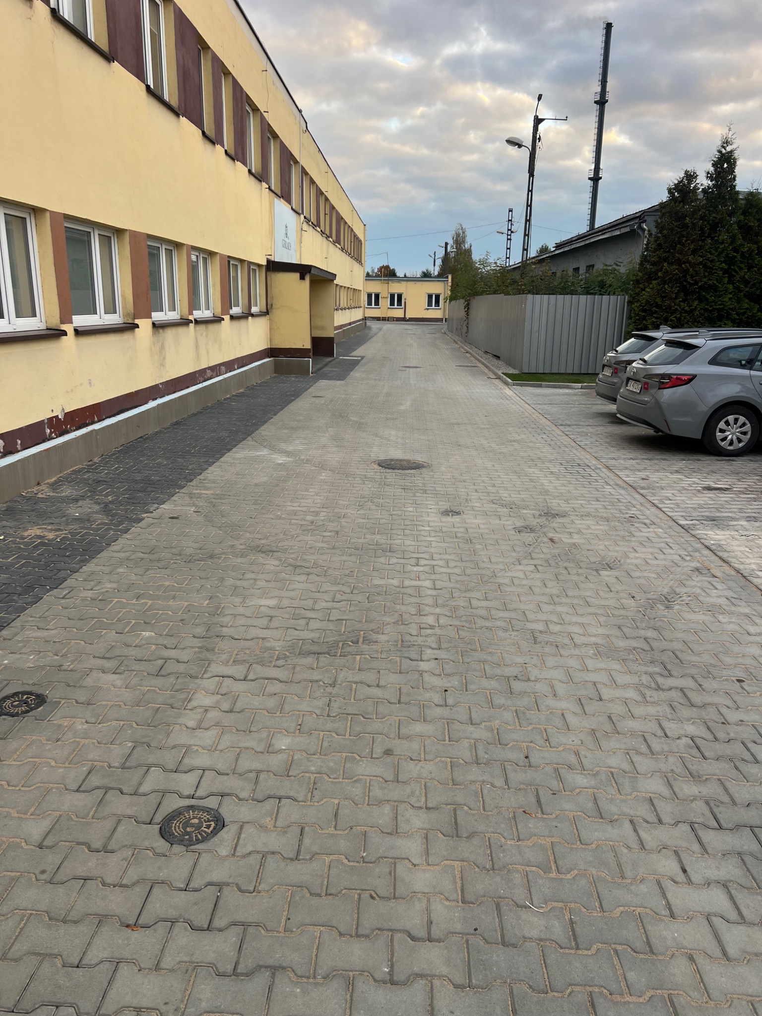 Szara kostka brukowa ułożona na długim podjeździe przed budynkiem z żółtą elewacją. W tle zaparkowane dwa samochody osobowe.