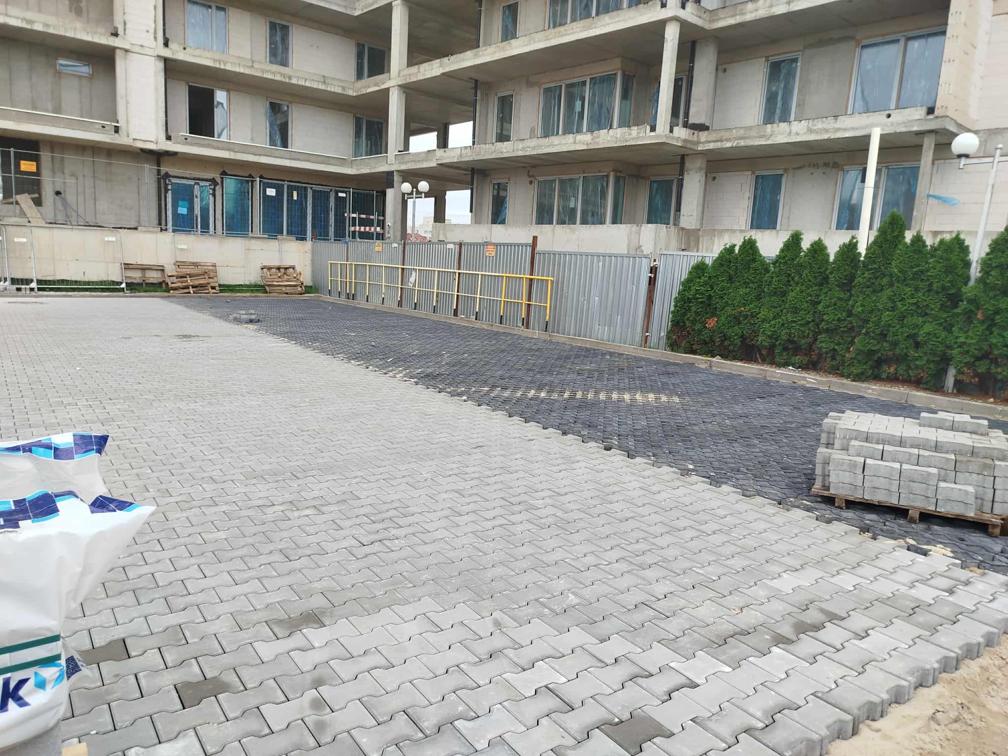 Częściowo ułożona, szara kostka brukowa na tle surowego budynku wielorodzinnego. Widoczne palety z kostką i fragmenty czarnej kostki. Prace brukarskie w toku.
