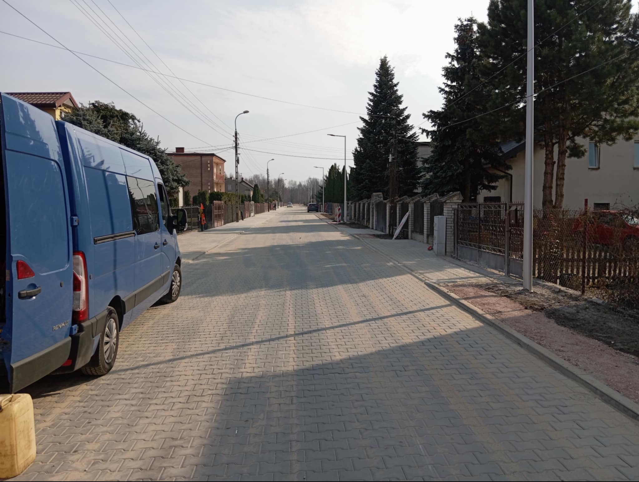 Ulica z nową, szarą kostką brukową ułożoną w jodełkę. Po lewej stronie zaparkowany niebieski bus z otwartymi drzwiami. W tle domy i drzewa.
