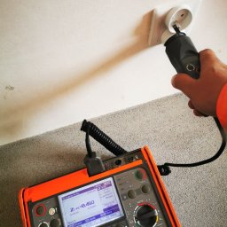 Fhu Tech-Instal Mariusz Zygnerski - Pomiar gniazdka elektrycznego miernikiem Sonel MPI-530. Widoczna dłoń w pomarańczowym rękawie i wynik pomiaru impedancji pętli zwarcia na wyświetlaczu.