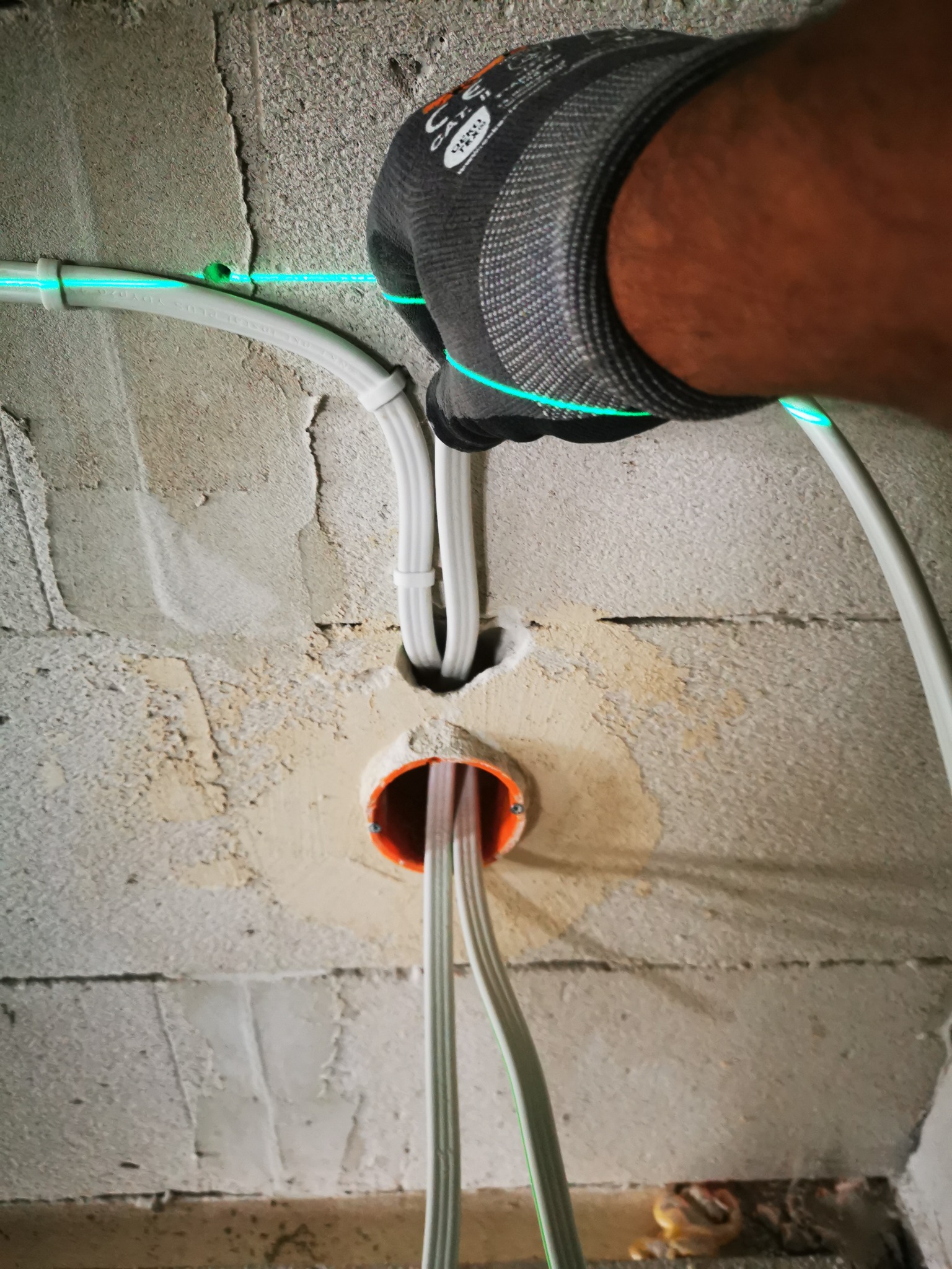 Białe przewody w ścianie, prowadzone przez pomarańczową puszkę instalacyjną. Ręka w rękawicy roboczej, zielona linia lasera wyznacza trasę kabli.