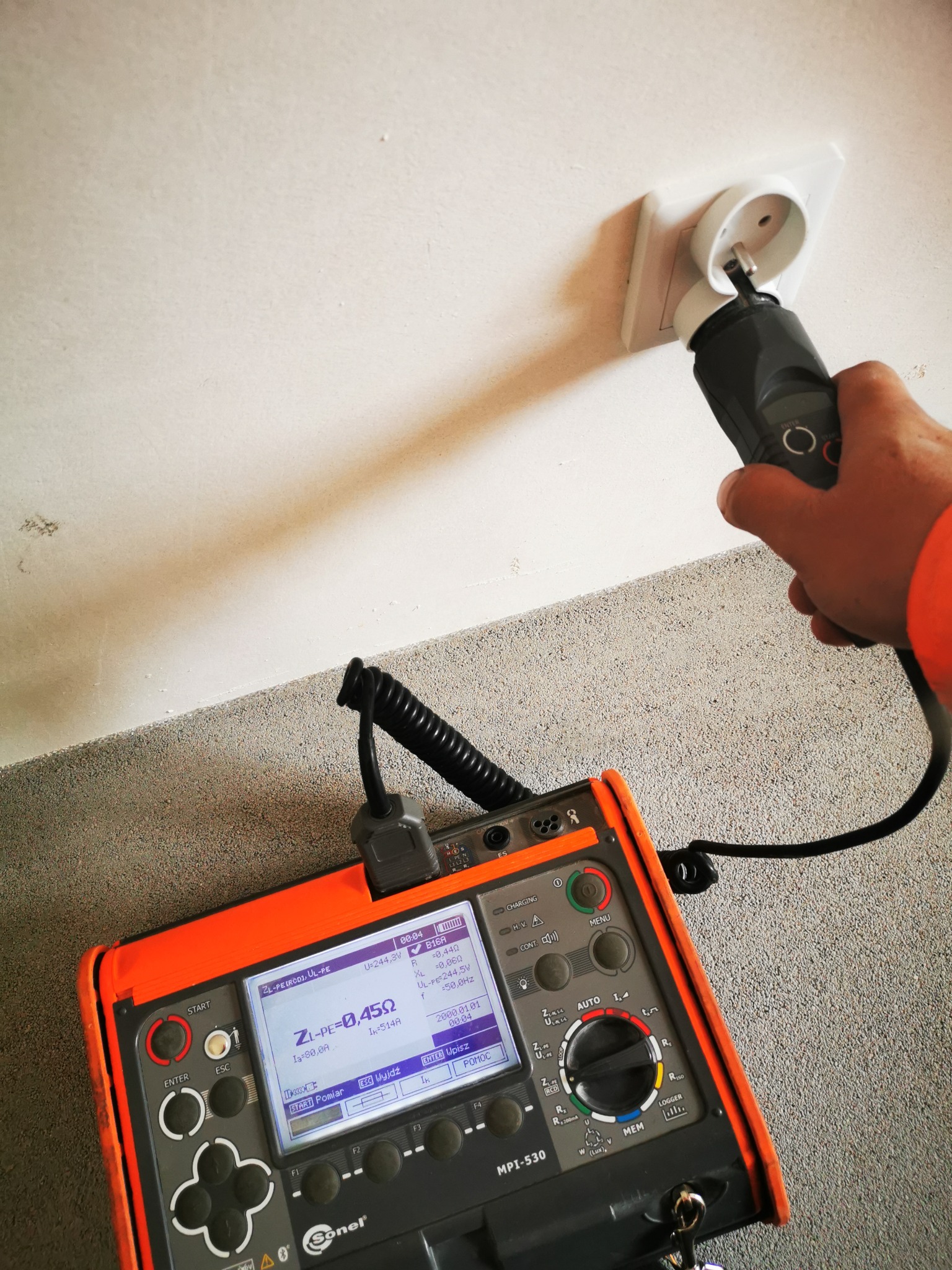 Pomiar gniazdka elektrycznego miernikiem Sonel MPI-530. Widoczna dłoń w pomarańczowym rękawie i wynik pomiaru impedancji pętli zwarcia na wyświetlaczu.