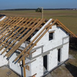 Usługi budowlane - Budowa domu z białych bloczków, widok z góry na konstrukcję dachu z drewnianymi belkami. W tle pola uprawne i błękitne niebo.