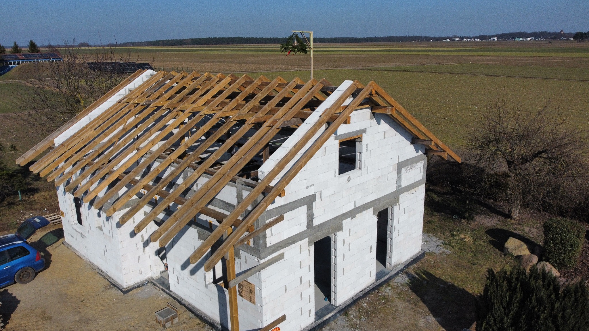 Budowa domu z białych bloczków, widok z góry na konstrukcję dachu z drewnianymi belkami. W tle pola uprawne i błękitne niebo.
