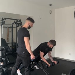 HealthStudio - Trener personalny nadzoruje ćwiczenie bicepsów z hantlami na ławce skośnej w siłowni. Widoczny sprzęt do ćwiczeń i lustro. Obaj mężczyźni ubrani na czarno.