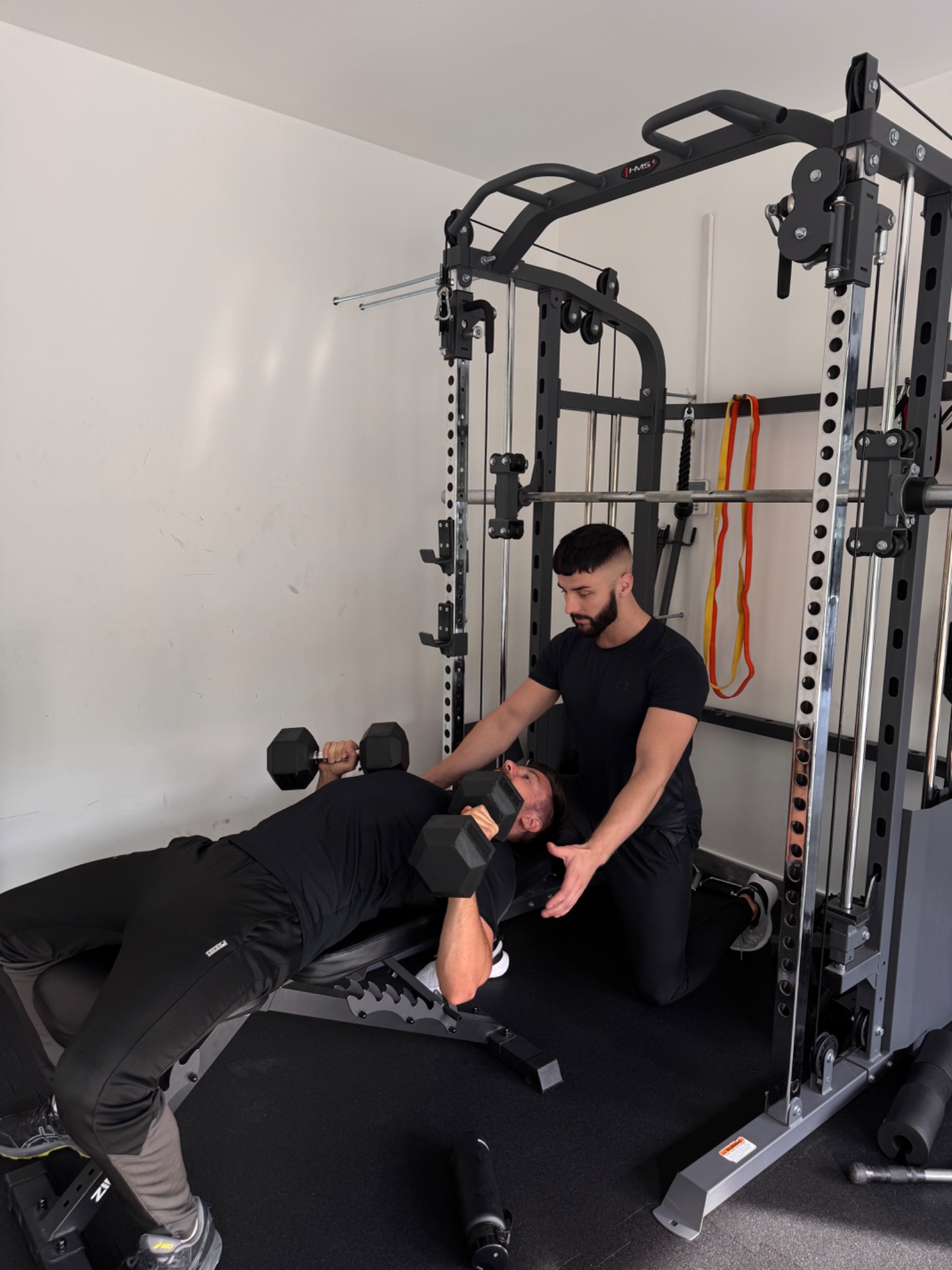 Trener asekuruje mężczyznę ćwiczącego wyciskanie hantli na ławce w siłowni. Sprzęt HMS, czarne ubrania, profesjonalne wsparcie, dbałość o technikę.