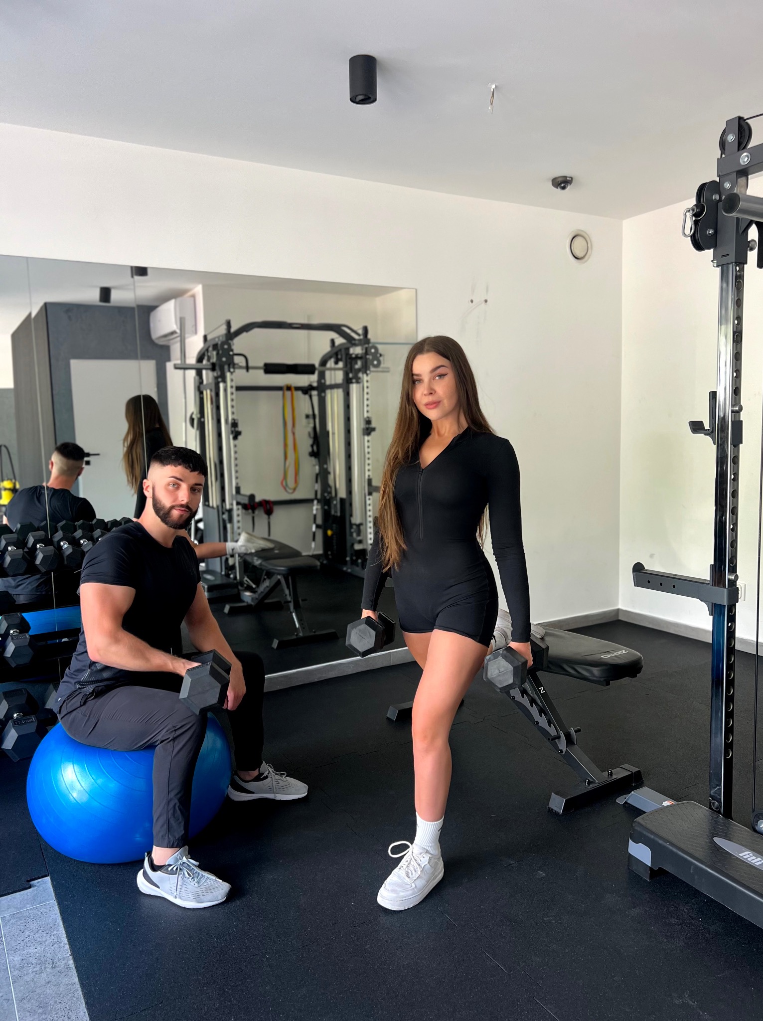 Para w czarnych strojach ćwiczy z hantlami w jasnej siłowni. Mężczyzna siedzi na niebieskiej piłce, kobieta stoi obok ławeczki do ćwiczeń.