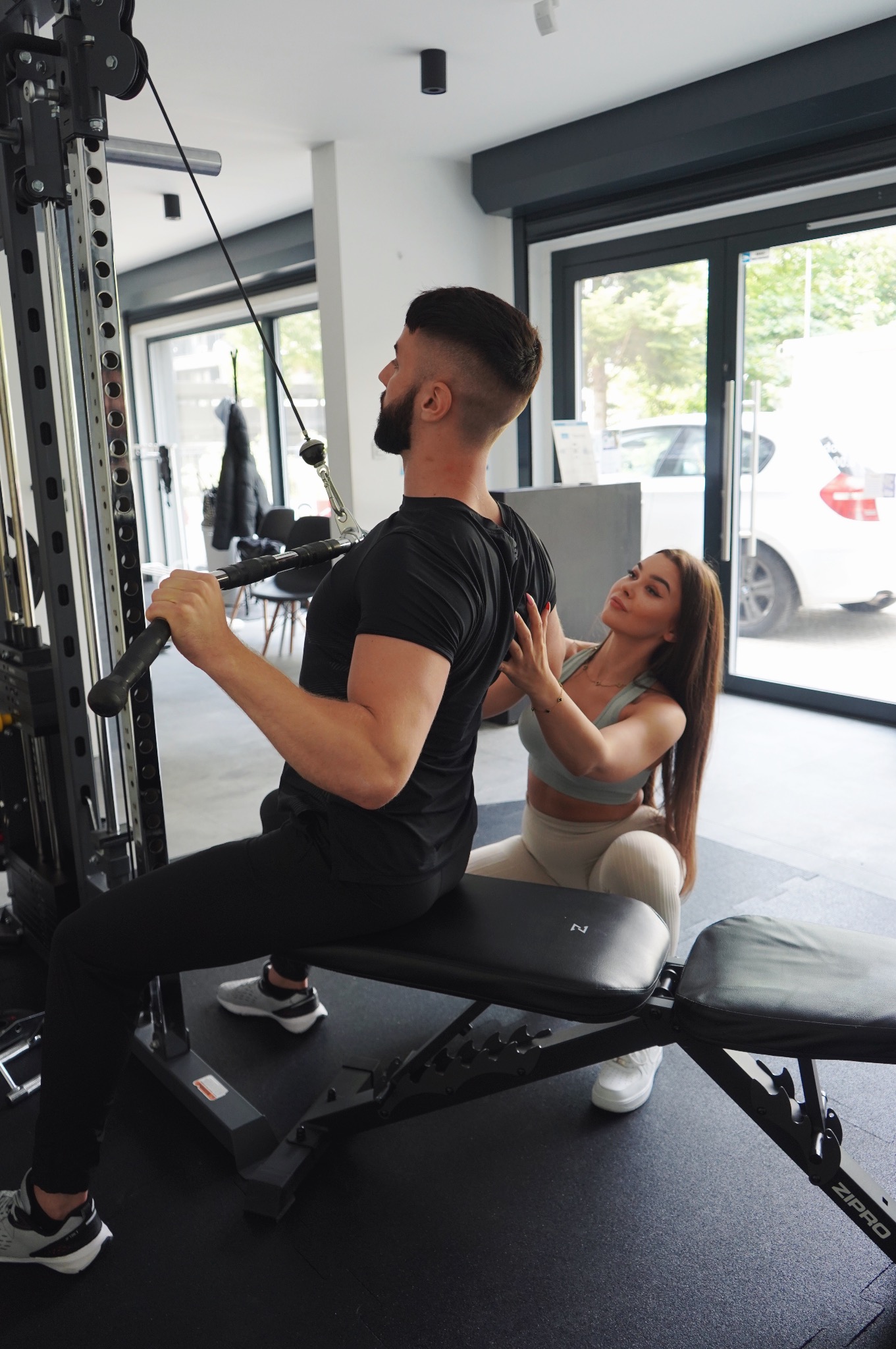 Mężczyzna ćwiczy na maszynie pod okiem trenerki w sportowym stroju. Kobieta koryguje jego postawę. Siłownia z dużymi oknami w tle.