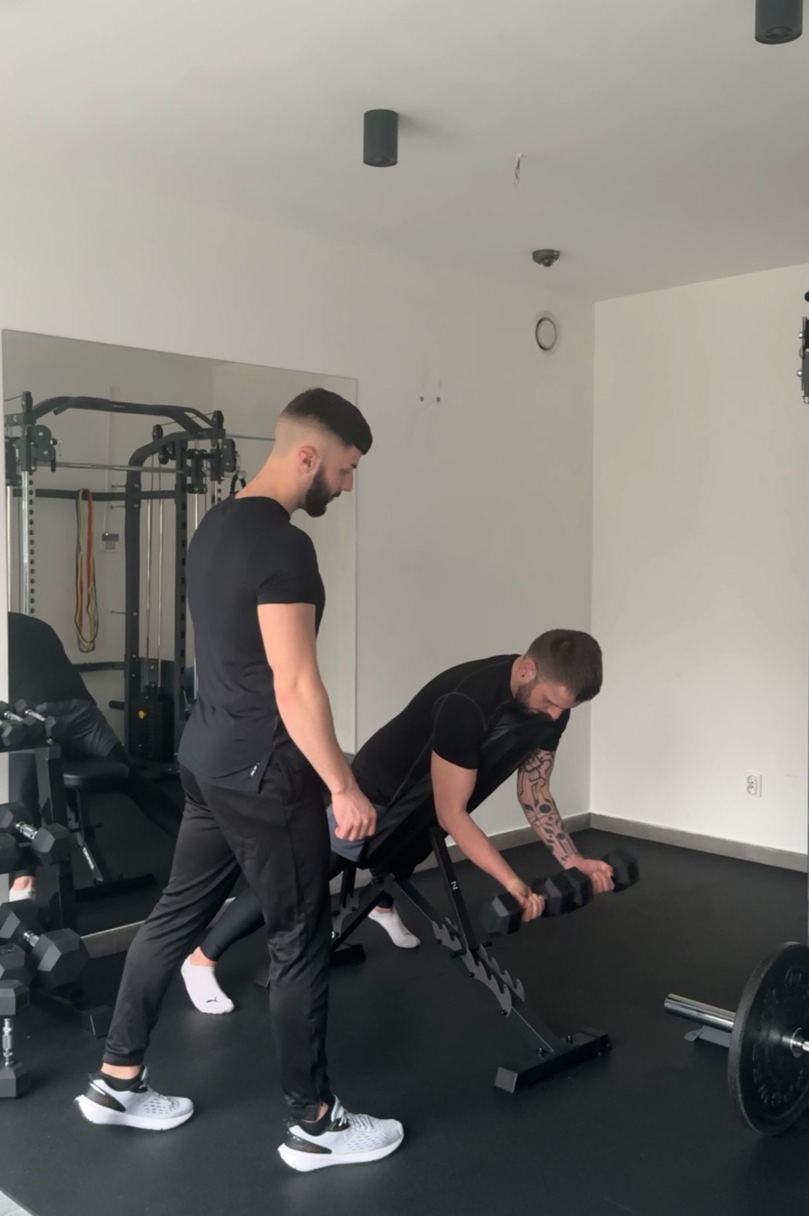 Trener personalny nadzoruje ćwiczenie bicepsów z hantlami na ławce skośnej w siłowni. Widoczny sprzęt do ćwiczeń i lustro. Obaj mężczyźni ubrani na czarno.