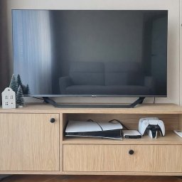 NaWarsztacie - Meble na wymiar - Jasna szafka RTV z drewna z czarnymi nóżkami i okrągłymi uchwytami, na której stoi telewizor. Na szafce dekoracje świąteczne i konsola do gier.