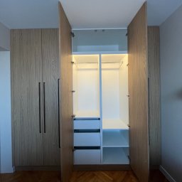 NaWarsztacie - Meble na wymiar - Jasna szafa wnękowa z drewnianymi frontami i czarnymi uchwytami, otwarte drzwi ukazują białe wnętrze z półkami i szufladami, oświetlenie LED wewnątrz.