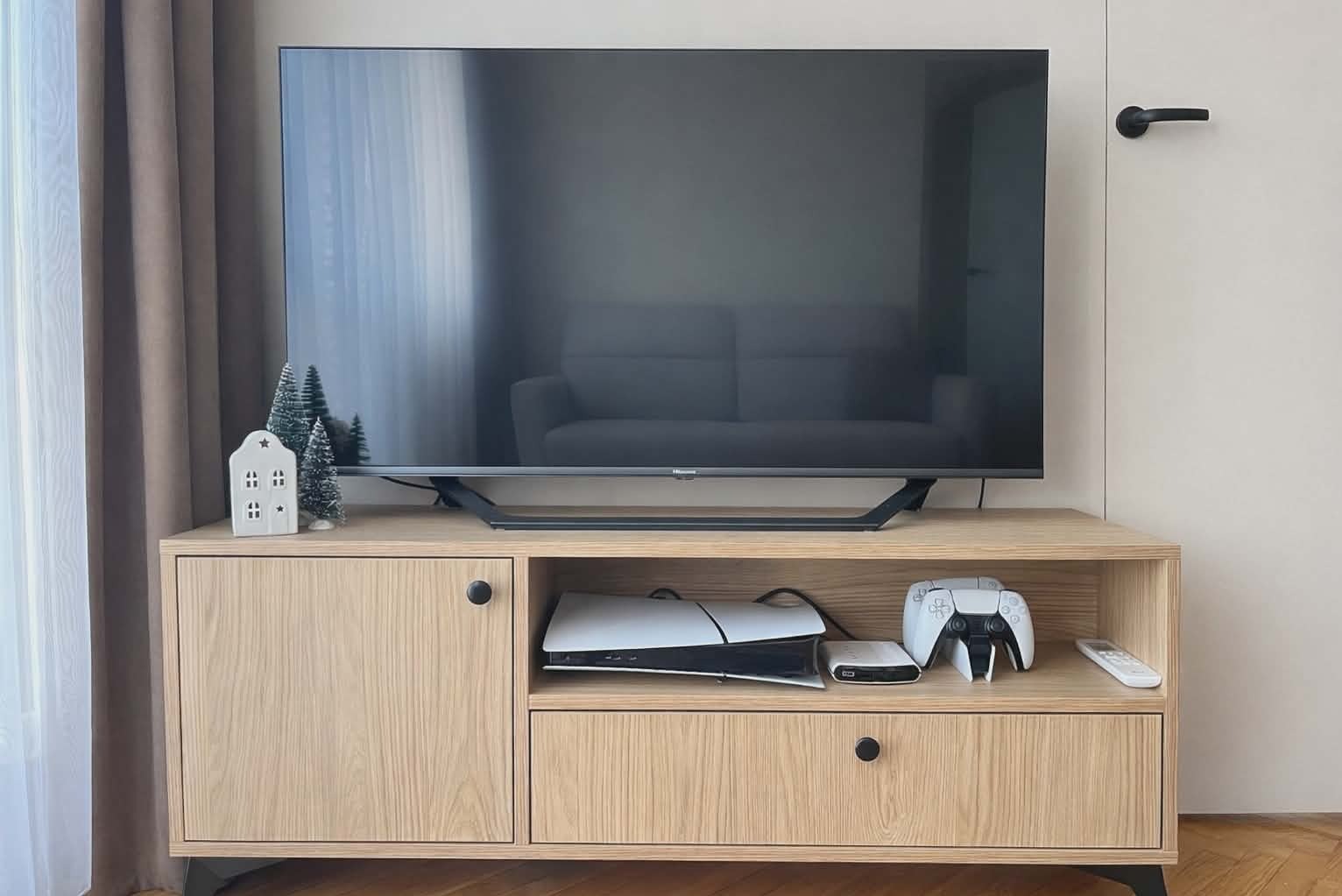 Jasna szafka RTV z drewna z czarnymi nóżkami i okrągłymi uchwytami, na której stoi telewizor. Na szafce dekoracje świąteczne i konsola do gier.