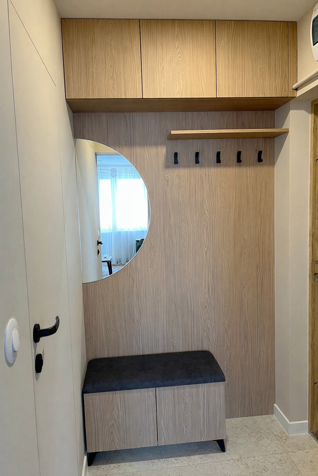 Funkcjonalna zabudowa przedpokoju z szafkami górnymi, wieszakami i siedziskiem z lustrem w kształcie półksiężyca. Jasne drewno z wyraźnym usłojeniem.