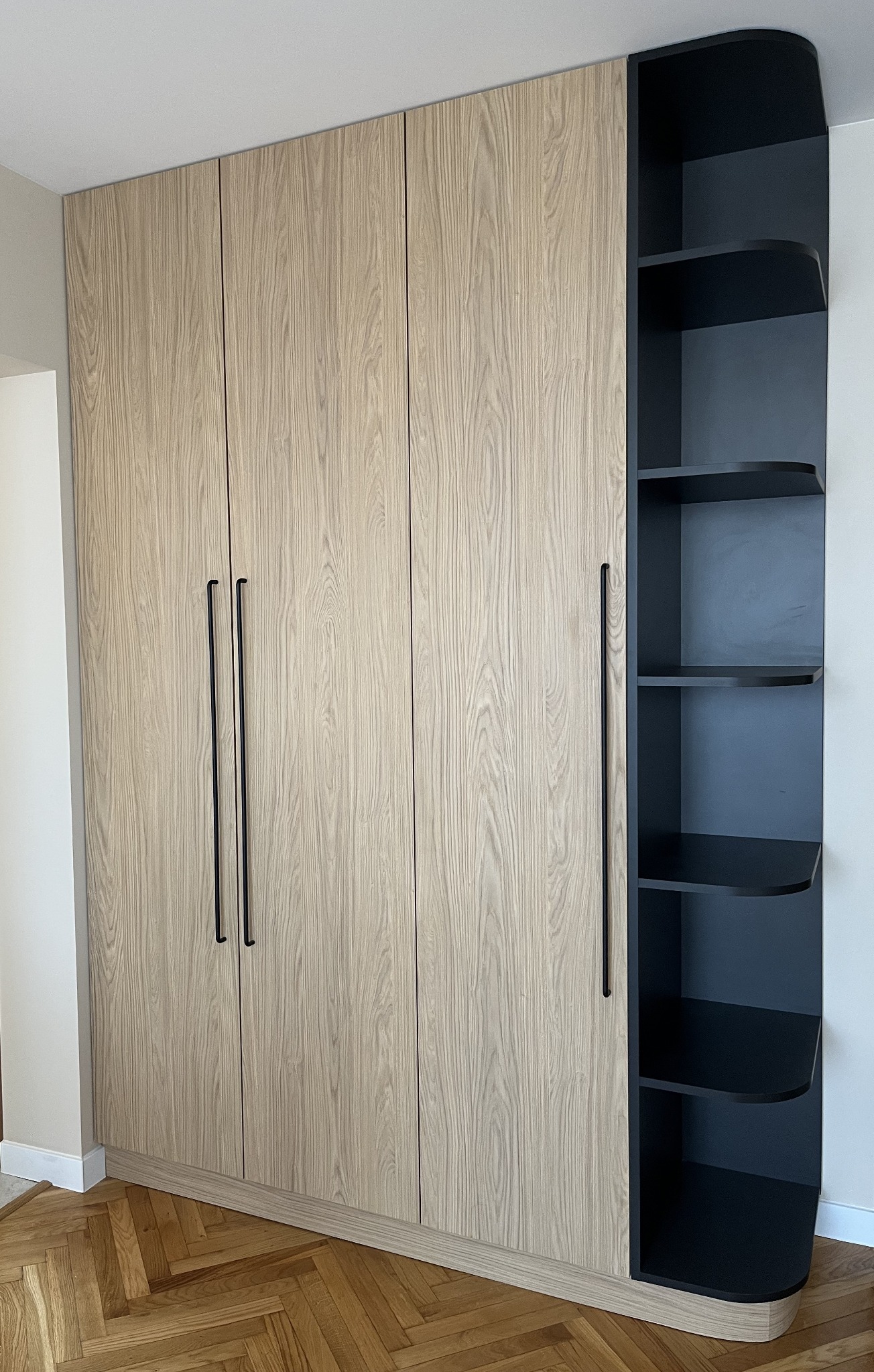 Jasna szafa na wymiar z czterema drzwiami i czarnym regałem z półkami po prawej stronie, na tle parkietu ułożonego w jodełkę.