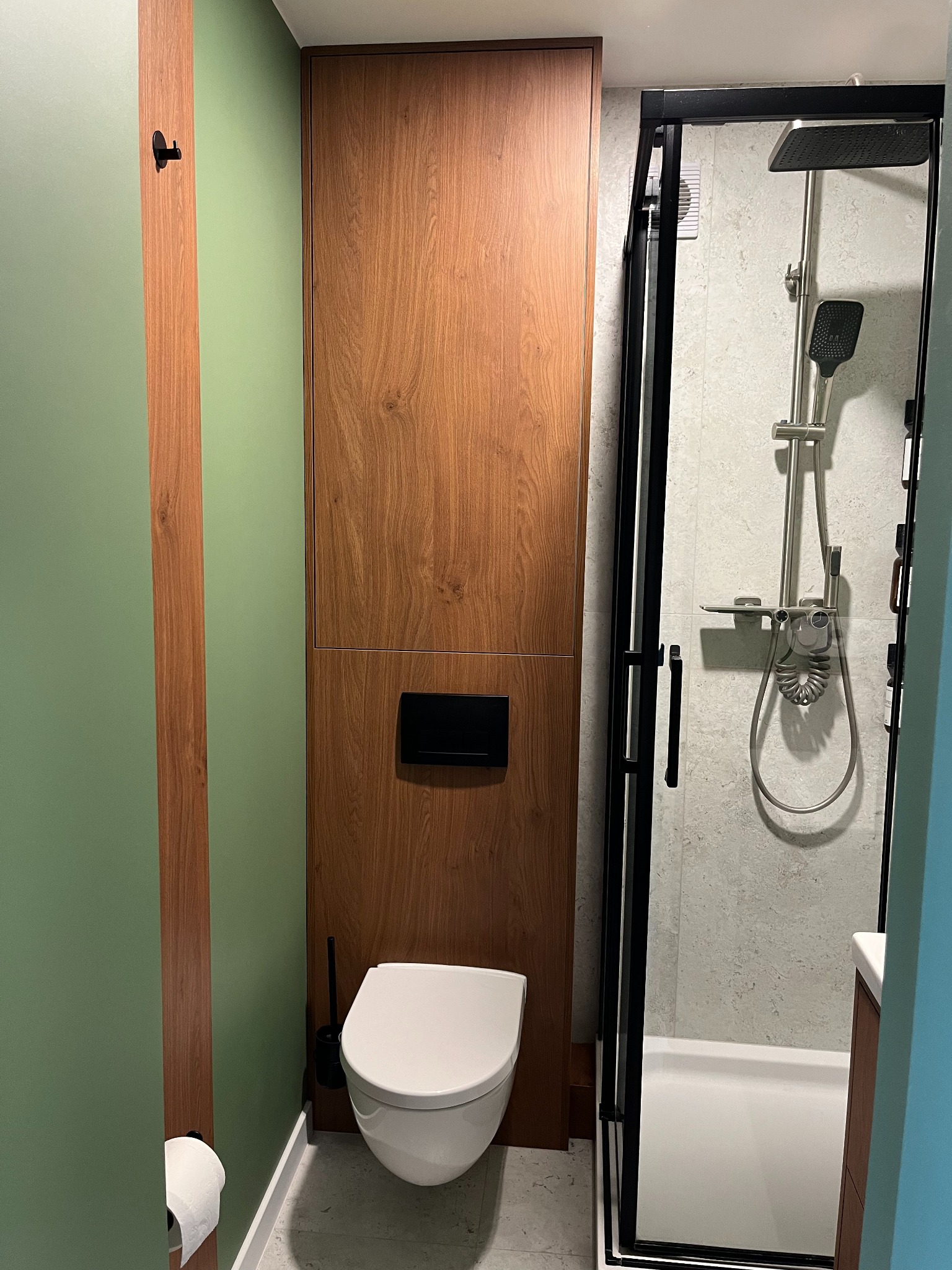 Nowoczesna łazienka z drewnianą zabudową toalety i kabiną prysznicową z czarnymi ramami. Minimalistyczny design, zielona ściana z drewnianym akcentem.