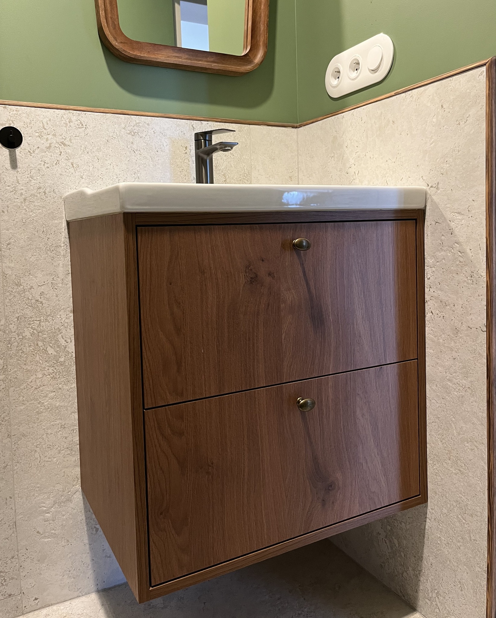 Drewniana szafka pod umywalkę z dwoma szufladami i okrągłymi uchwytami w łazience z zielonymi ścianami i lustrem w drewnianej ramie.