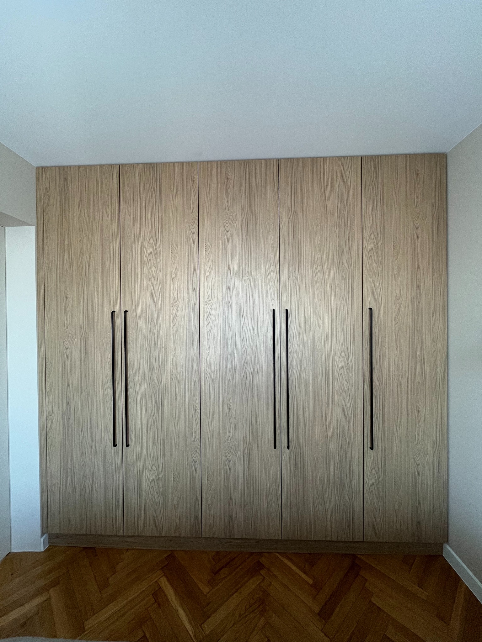 Szafa wnękowa z jasnego drewna z sześcioma drzwiami i długimi, czarnymi uchwytami, na tle parkietu ułożonego w jodełkę.