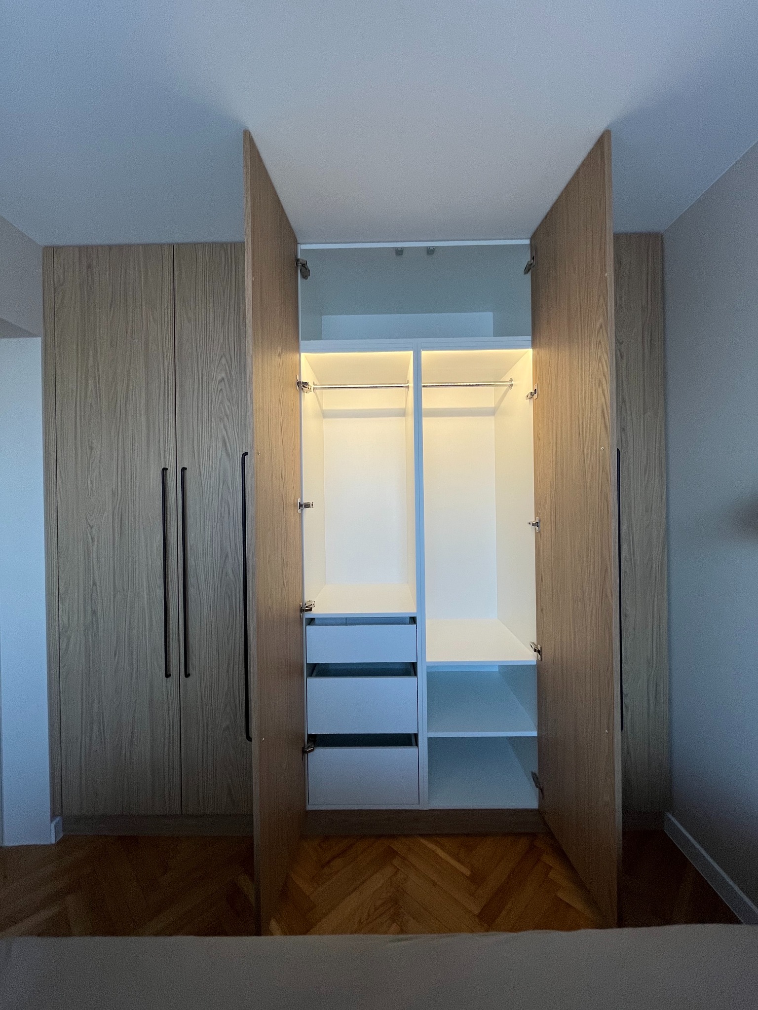 Jasna szafa wnękowa z drewnianymi frontami i czarnymi uchwytami, otwarte drzwi ukazują białe wnętrze z półkami i szufladami, oświetlenie LED wewnątrz.