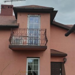 Fenix - Dwupiętrowy dom z balkonem i nowymi oknami PCV. Widoczna osoba sprzątająca teren wokół budynku. Elewacja w odcieniach różu, dachówka brązowa. Antena na dachu.