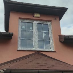 Fenix - Nowe, białe okna z szybami w kratkę, zamontowane w różowej elewacji domu z brązowym dachem. Widok z dołu, pochmurne niebo w tle. Okna zabezpieczone folią.