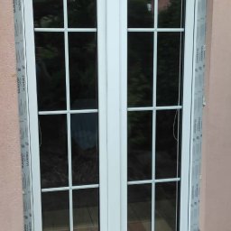 Fenix - Białe, dwuskrzydłowe okno PCV z podziałami szprosowymi, zamontowane w różowej ścianie. Widoczny karton z napisem 'Fragile'.