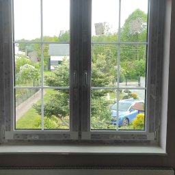 Fenix - Białe okno PCV z widokiem na ogród i niebieski samochód. Nowe okno z szybami zespolonymi i białymi ramami. Widoczny grzejnik pod oknem.