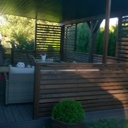 Tomasz Brodowski - Zadaszony taras z drewnianą konstrukcją i meblami ogrodowymi, otoczony zielenią. Widoczne detale wykończenia i elementy dekoracyjne.