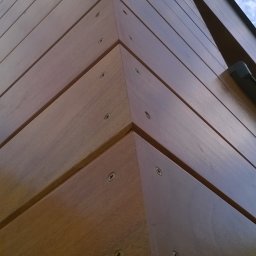 Tomasz Brodowski - Kąt budynku obłożonego drewnianymi panelami, mocowanymi na śruby. Widoczne niebo w tle. Ujęcie z dołu, podkreślające geometrię i fakturę drewna.