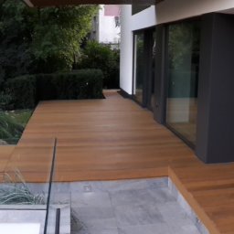 Tomasz Brodowski - Nowoczesny taras drewniany z jasnego drewna, otoczony zielenią i szklaną balustradą, przylegający do budynku z dużymi oknami.