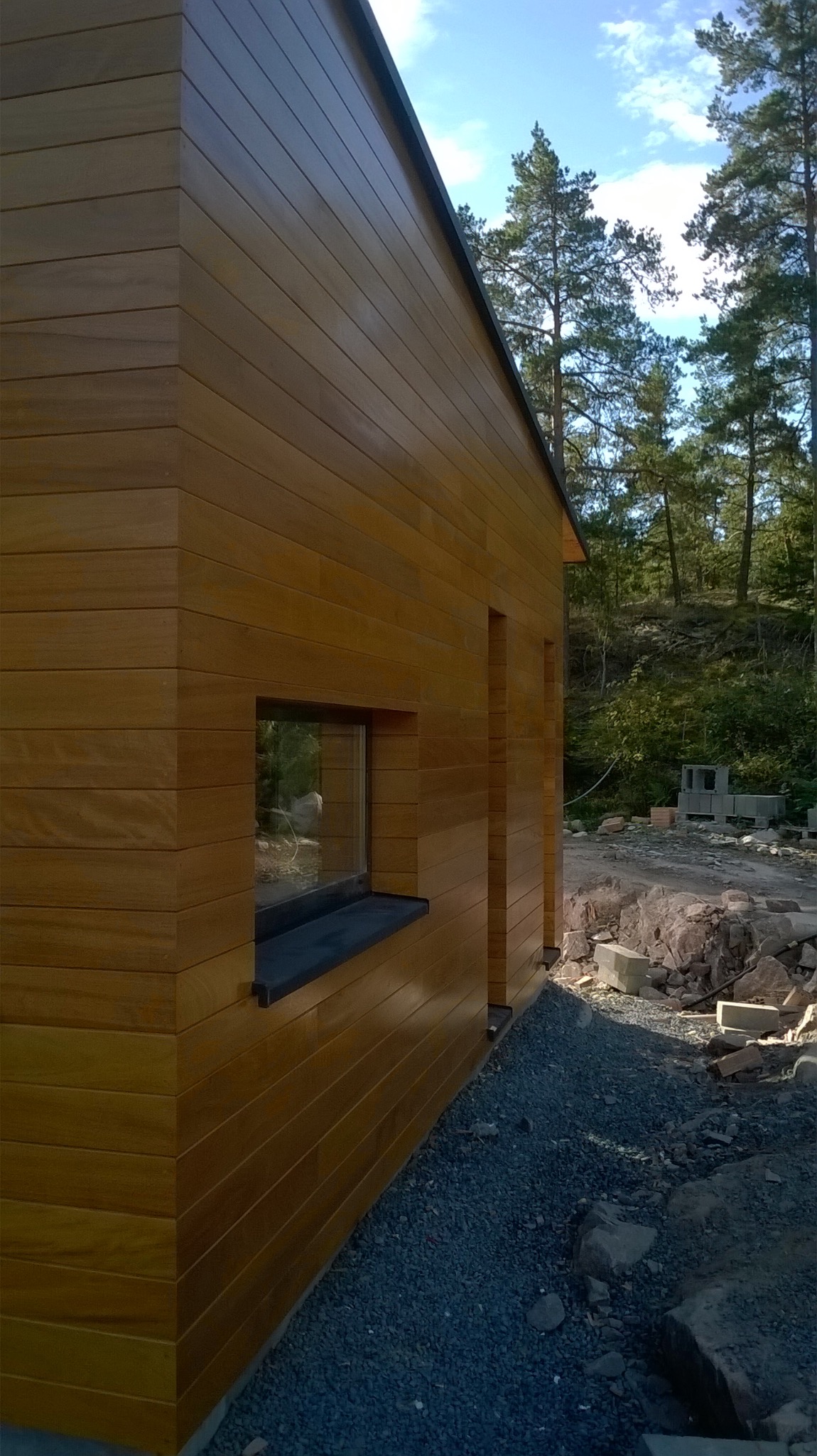 Elewacja domu z drewnianą okładziną w Rokitkach. Widok z boku, okno z czarnym parapetem, szara podsypka. W tle drzewa i teren w trakcie prac wykończeniowych.