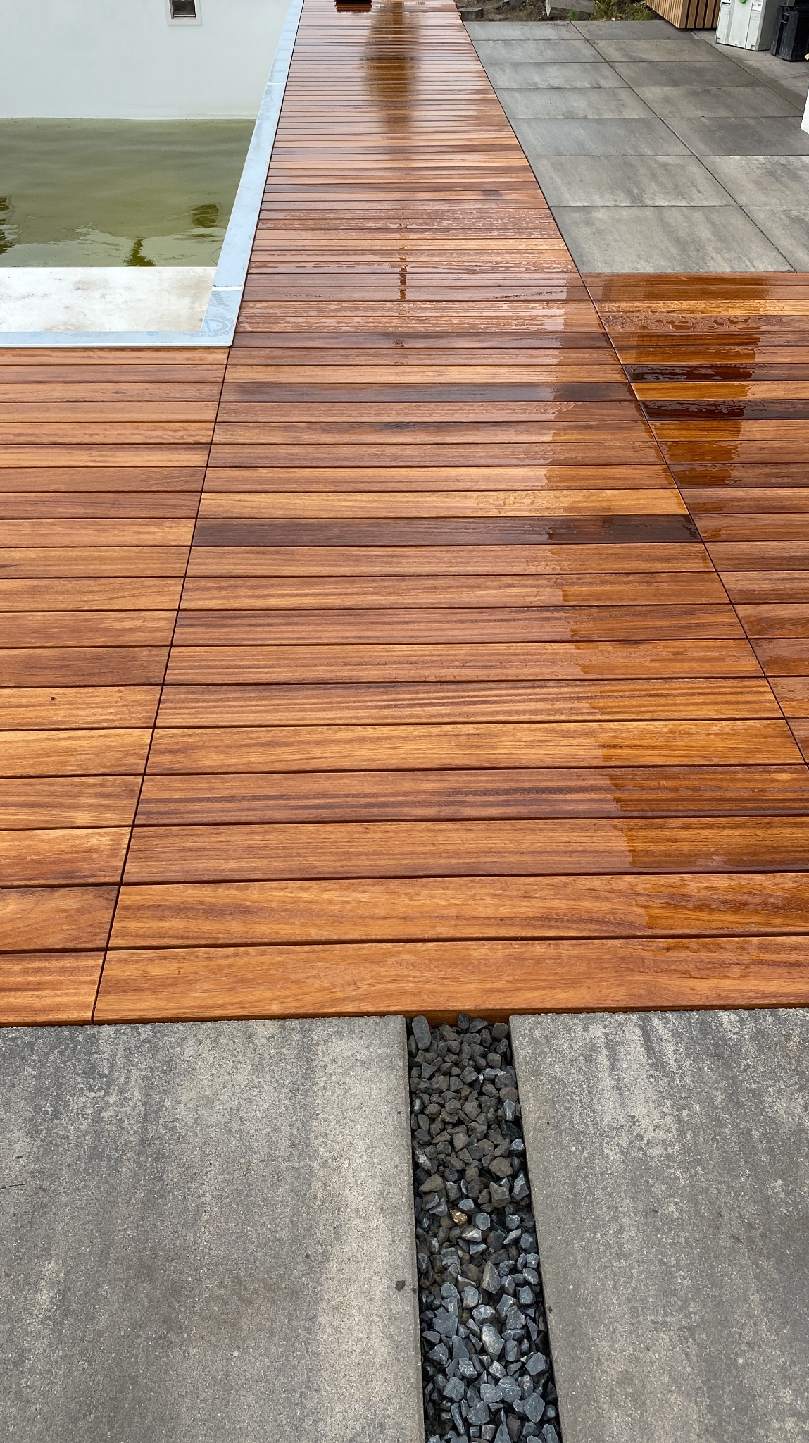 Lśniący, drewniany taras z desek ułożonych równolegle, przylegający do basenu i szarej nawierzchni z kamiennym drenażem. Widok z góry, podkreślający detale drewna.