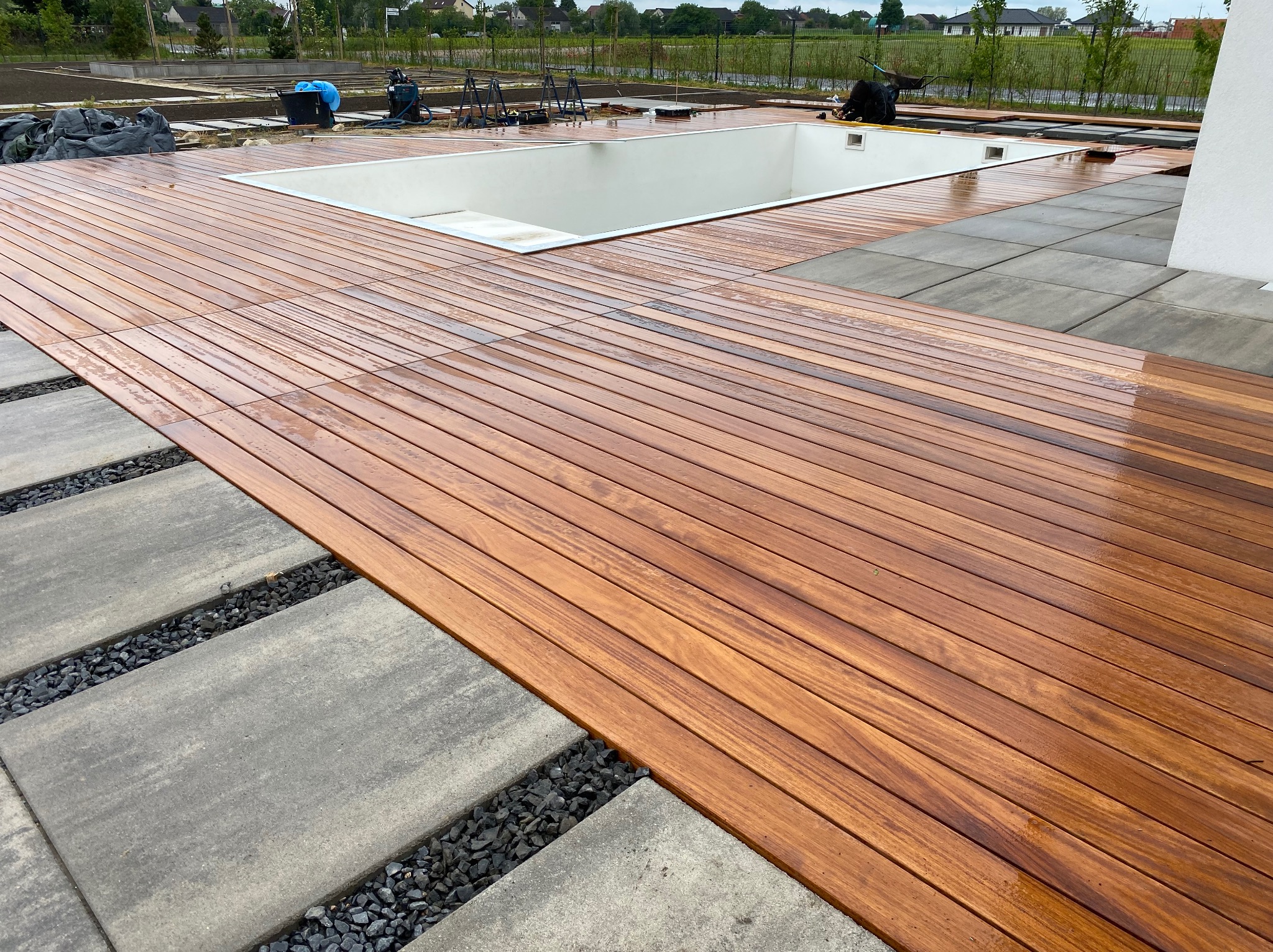 Nowoczesny taras z drewna egzotycznego otaczający basen, z elementami z szarych płyt betonowych i czarnego grysu. Widok na ogród i domy w tle.