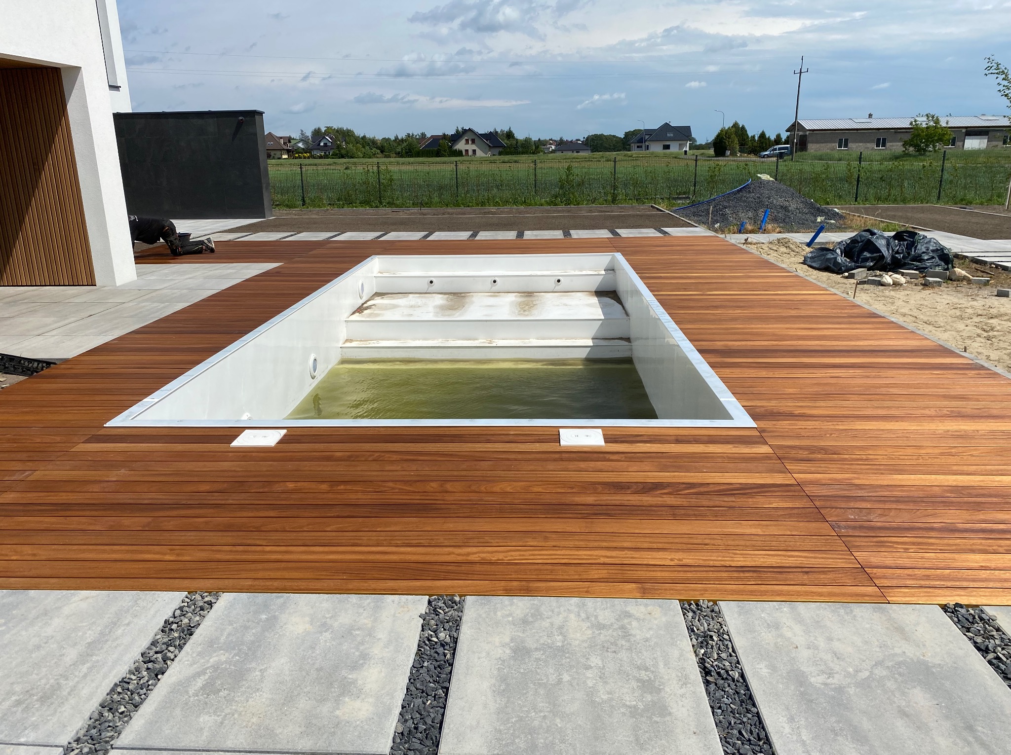 Nowoczesny taras z drewna egzotycznego otaczający basen, z elementami betonowymi i kamiennymi. Widok na ogród i domy w tle, w słoneczny dzień.