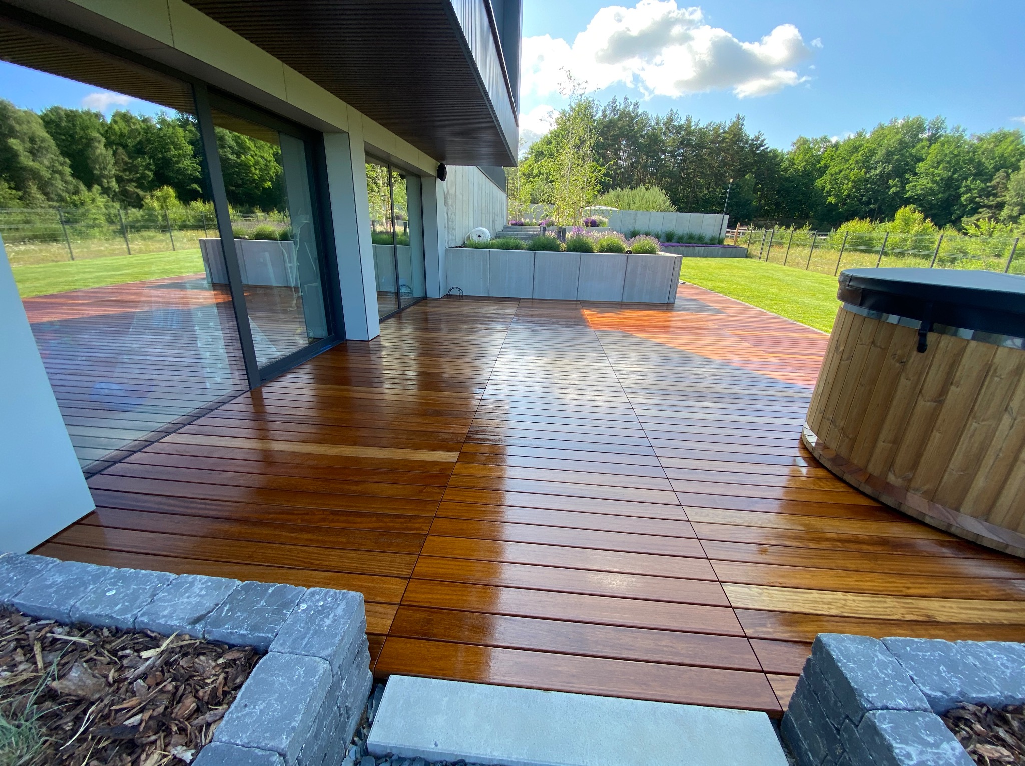 Nowoczesny taras z drewna egzotycznego, z jacuzzi i elementami kamiennymi. Widok na zadbany ogród i nowoczesny dom z dużymi przeszkleniami. Słoneczny dzień.