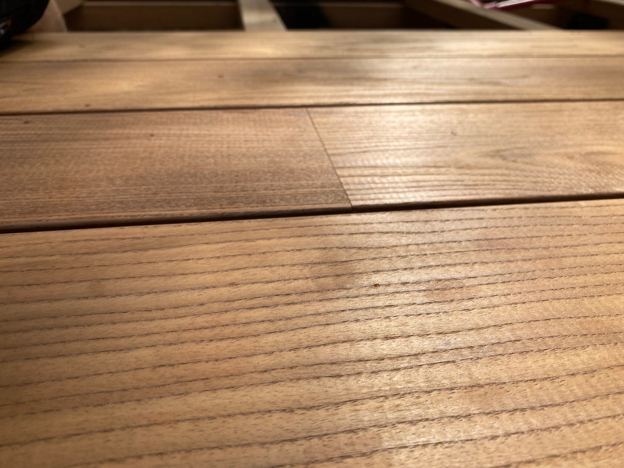 Jasnobrązowy taras drewniany z widoczną strukturą drewna i elementami konstrukcyjnymi w tle, wykonany z desek ułożonych równolegle.