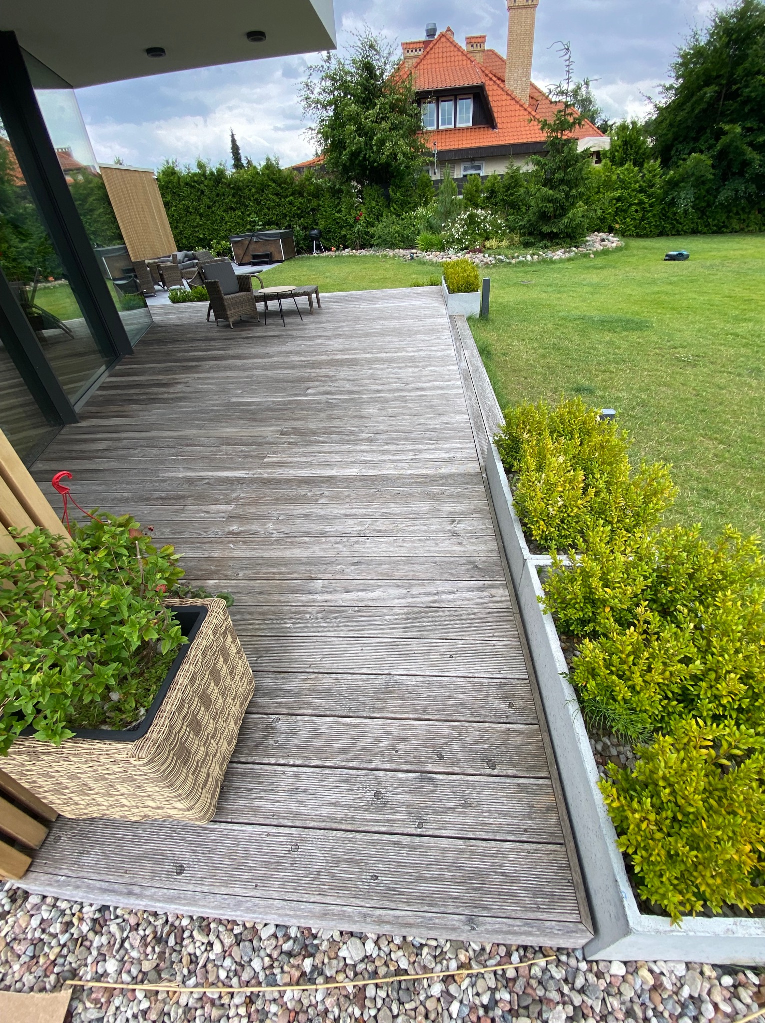 Szara, drewniana deska tarasowa z donicą i zielenią, widok na ogród z domem w tle. Minimalistyczny design, naturalne materiały, nowoczesna architektura.