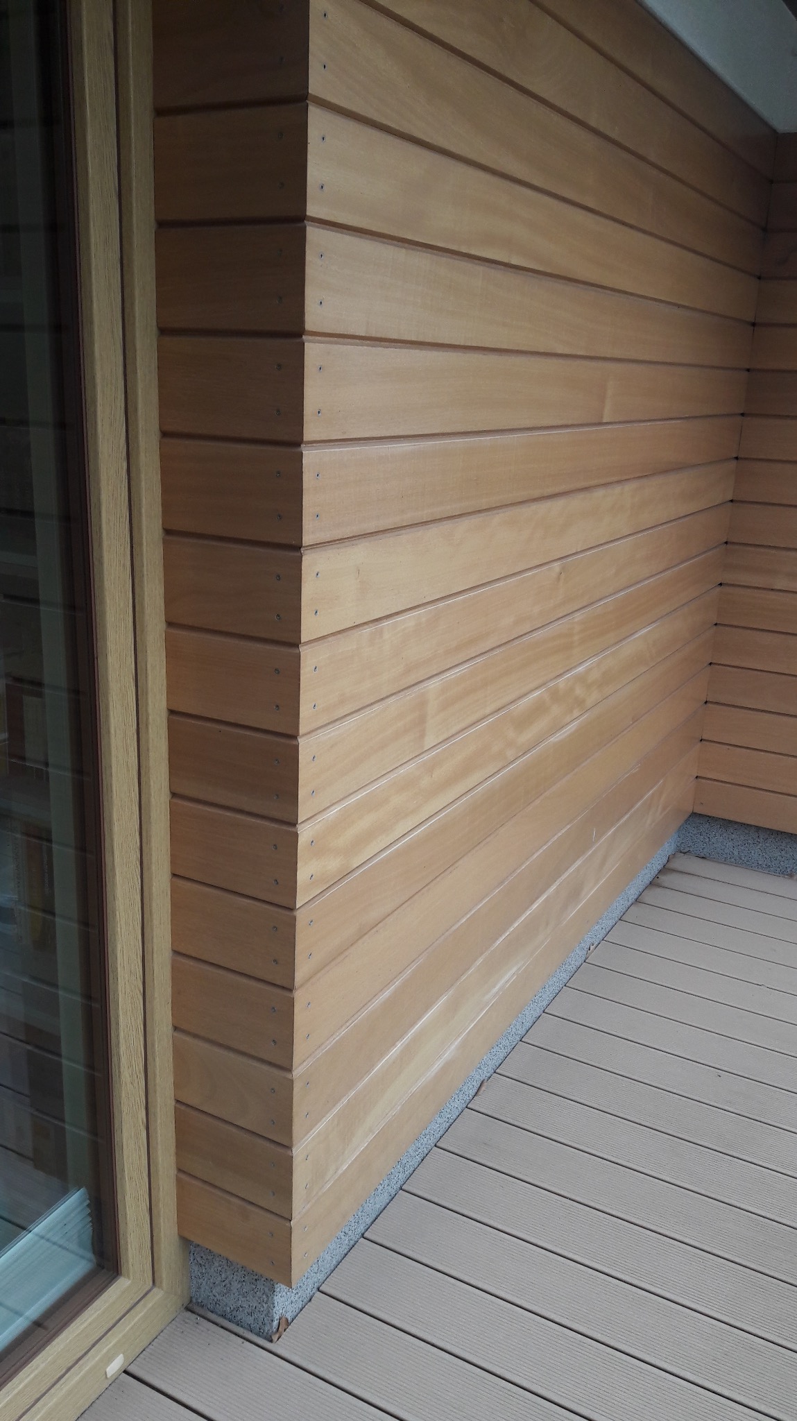 Jasne, drewniane deski elewacyjne i podłoga tarasu kompozytowego, widok z narożnika przy oknie. Detal wykończenia z widocznymi śrubami.