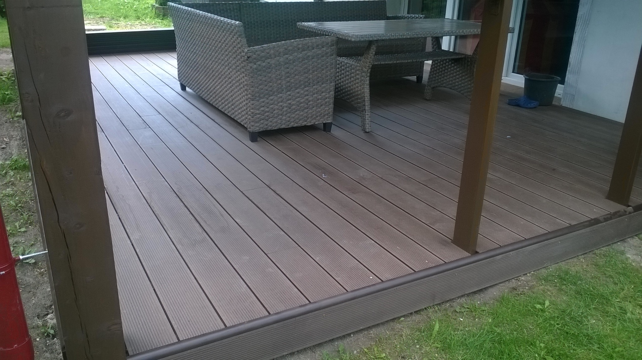 Kompozytowy taras z meblami ogrodowymi w odcieniach szarości, widok z niskiej perspektywy, z elementami zieleni w tle. Deski ułożone równolegle, widoczne słupki konstrukcyjne.