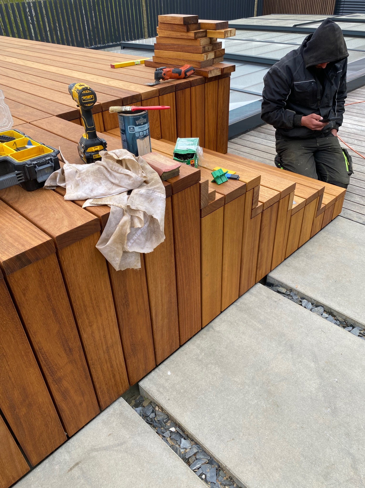 Montaż tarasu z drewna egzotycznego: deski, narzędzia, impregnat. Pracownik w bluzie z kapturem sprawdza coś w telefonie. Widoczne betonowe płyty i kamień.