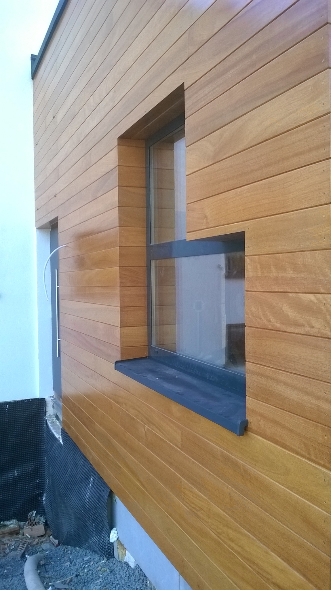 Elewacja budynku wykończona poziomymi deskami z jasnego drewna, widoczne okno z ciemną ramą i fragment drzwi. U dołu widoczna warstwa izolacyjna i gruz.