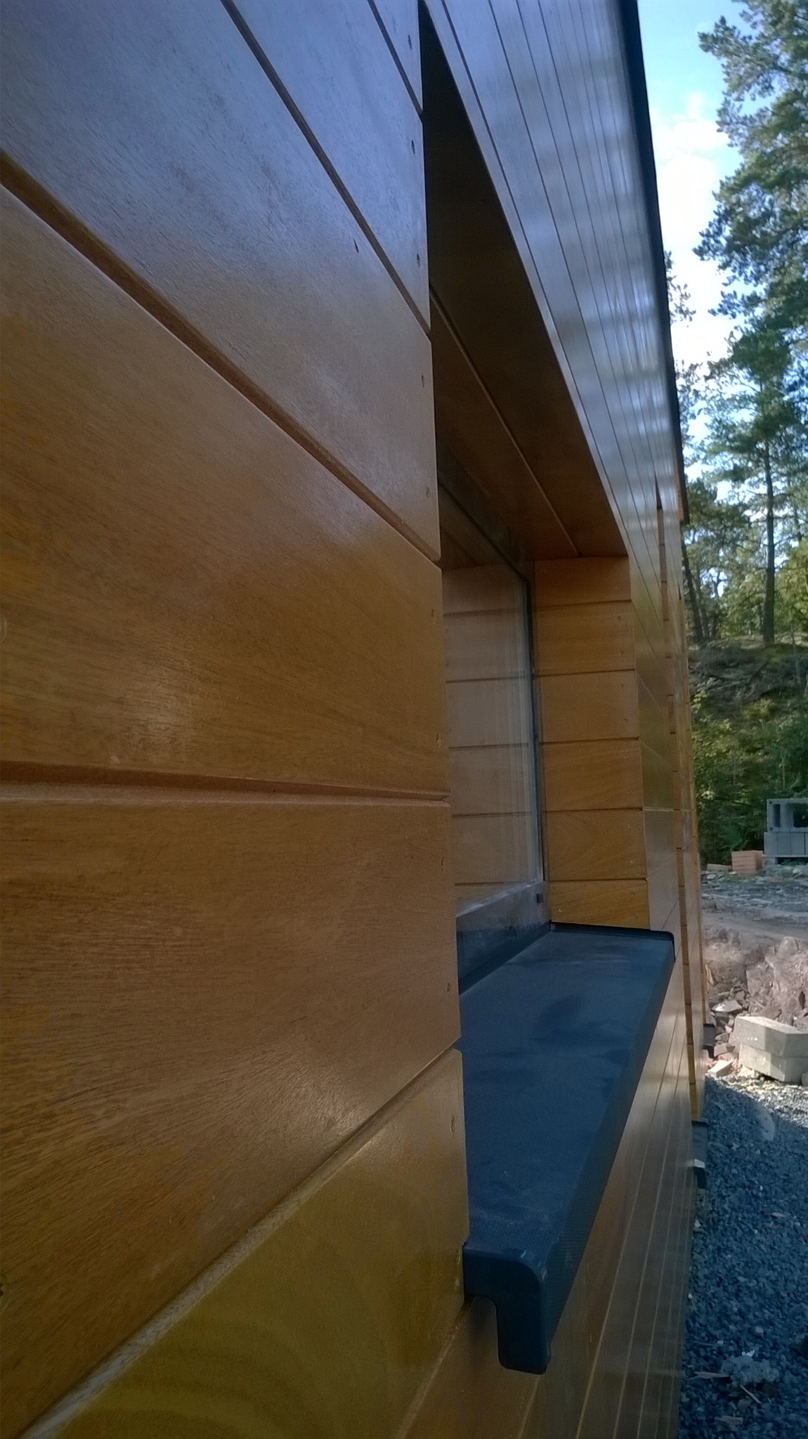 Detal drewnianej elewacji budynku z oknem i czarnym parapetem, widok z dołu pod kątem. W tle drzewa i elementy budowlane.