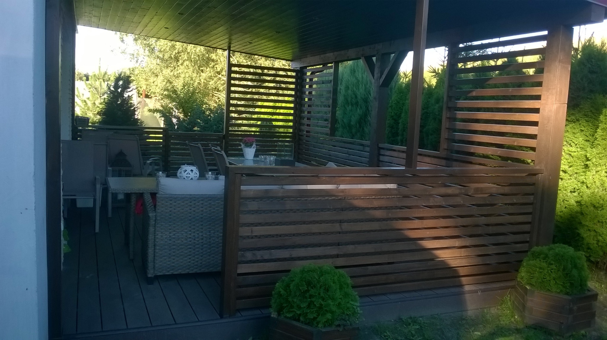 Zadaszony taras z drewnianą konstrukcją i meblami ogrodowymi, otoczony zielenią. Widoczne detale wykończenia i elementy dekoracyjne.