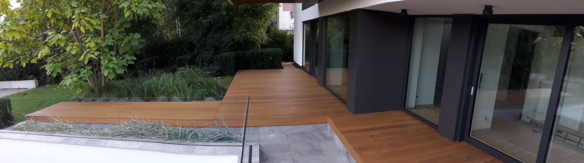 Nowoczesny taras drewniany z jasnego drewna, otoczony zielenią i szklaną balustradą, przylegający do budynku z dużymi oknami.