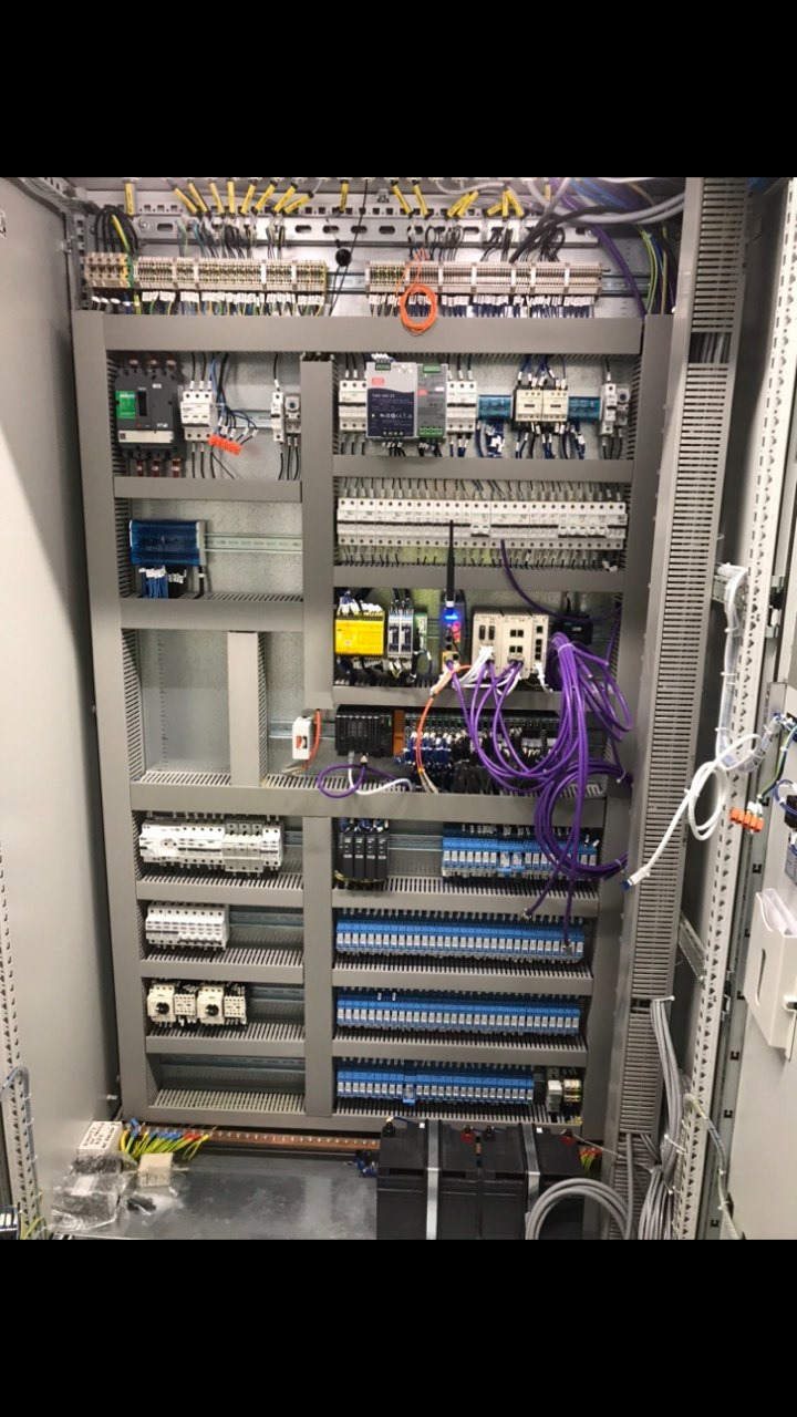 Szara szafa sterownicza z licznymi przewodami, bezpiecznikami i elementami elektronicznymi. Widoczne kable w kolorze fioletowym. Rozbudowana instalacja wewnątrz szafy.