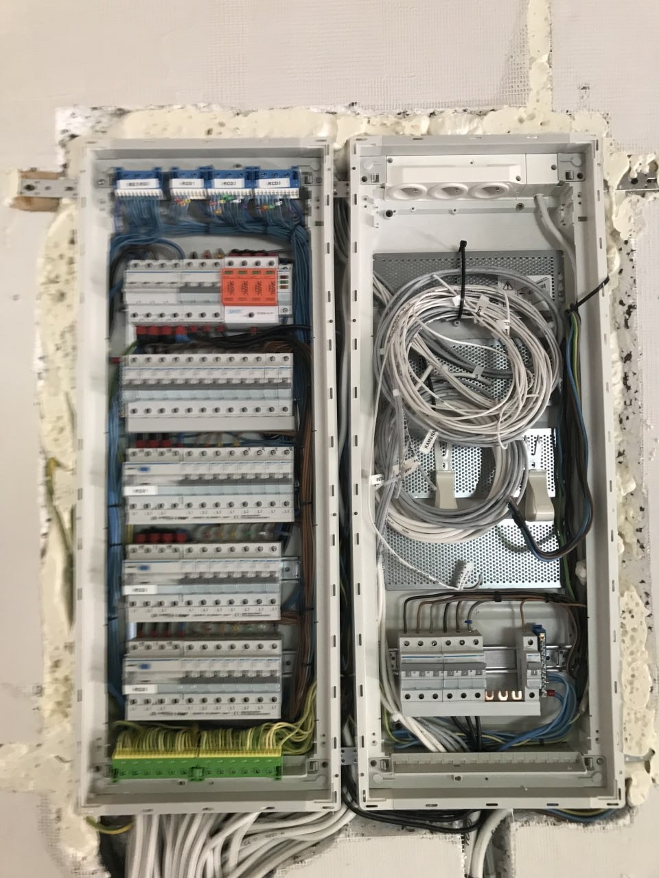 Otwarta, podwójna skrzynka elektryczna z licznymi przewodami i bezpiecznikami, zamontowana w ścianie z widocznymi śladami pianki montażowej. Układ przewodów uporządkowany.