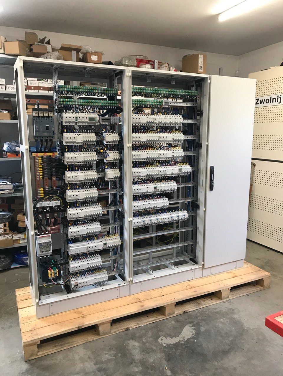 Rozbudowana szafa sterownicza z licznymi zabezpieczeniami i okablowaniem, umieszczona na palecie w pomieszczeniu warsztatowym. Widoczne elementy automatyki i zasilania.