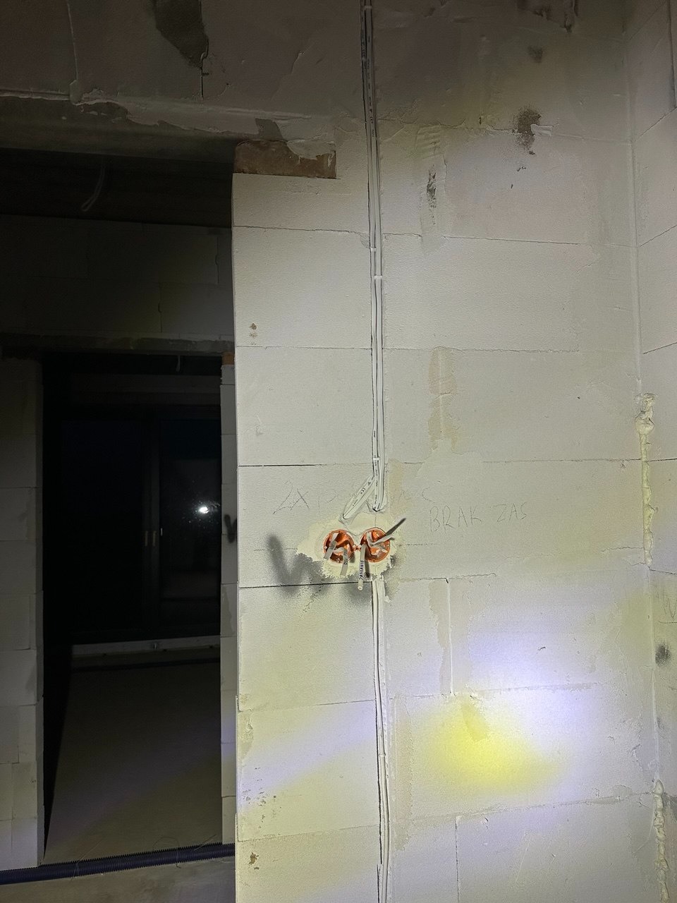 Wewnętrzna instalacja elektryczna w surowym wnętrzu: przewody w ścianie z bloczków, pomarańczowe puszki pod gniazdka, widoczne napisy na ścianie, surowy stan wykończenia.