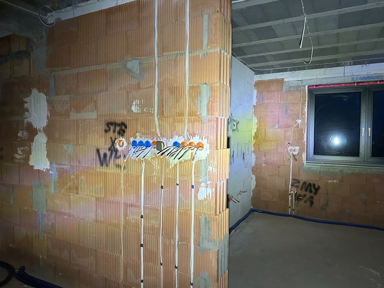 Surowa ściana z cegły z rozprowadzoną instalacją elektryczną: kable w peszlach, puszki pod gniazdka. Widoczne napisy na ścianie. W tle okno i fragment podłogi.