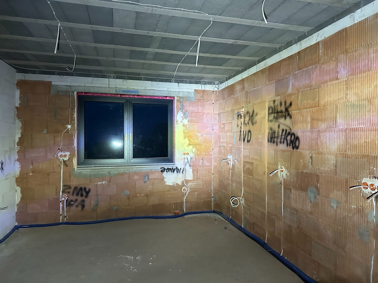 Surowy stan wnętrza z instalacją elektryczną w ścianach z cegły dziurawki. Widoczne kable, puszki elektryczne, okno i betonowa wylewka. Sufit z prefabrykatów.